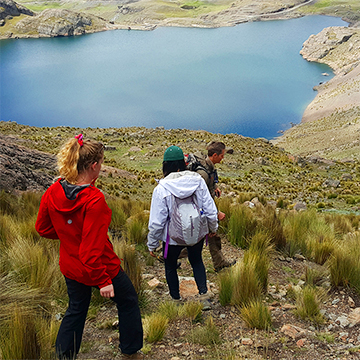 Three students hike down a hillside toward a bright blue lake