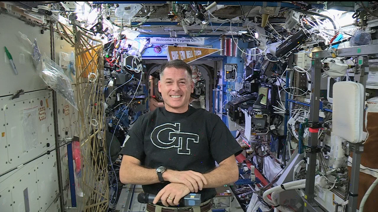 Screen shot of video chat with Shane Kimbrough on the International Space Station, wearing a GT logo shirt and with a "Give 'Em Hell Tech" pennant hanging behind him above some wires and equipment