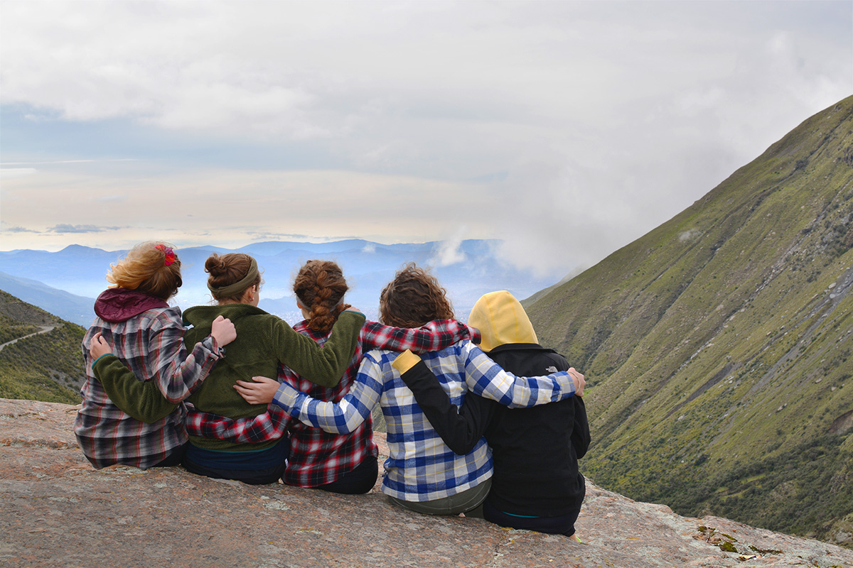 Five students sitting on a cliffside looking over the mountains with their arms around each other