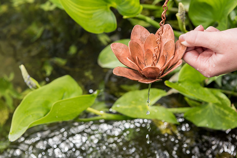 A lotus flower made of copper, in a leafy green setting over a body of water, with water dripping off of it