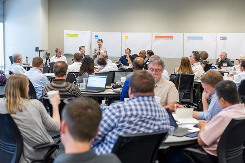 A design charette, or meeting: dozens of people seated at round tables, listening to a woman with a microphone speaking at the front of the room