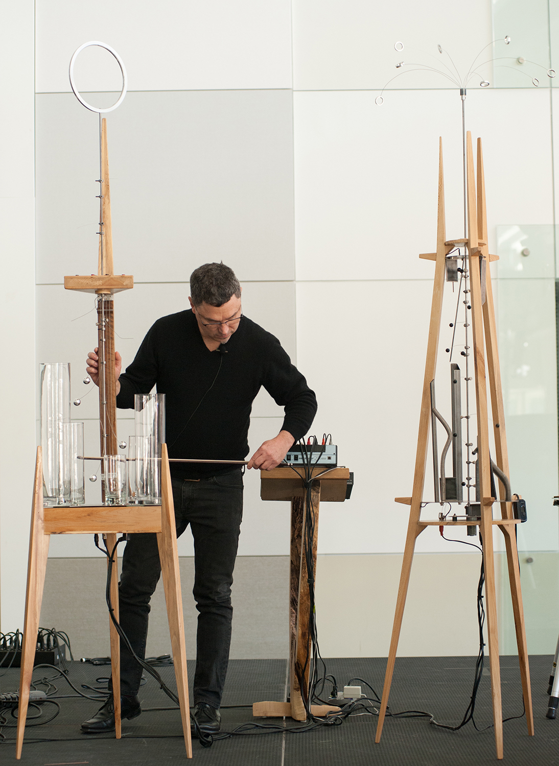 Man using a rod to make music on two easel-like wood towers that hold small metallic pieces and glass cylinders, and are connected by wires to an amplifier