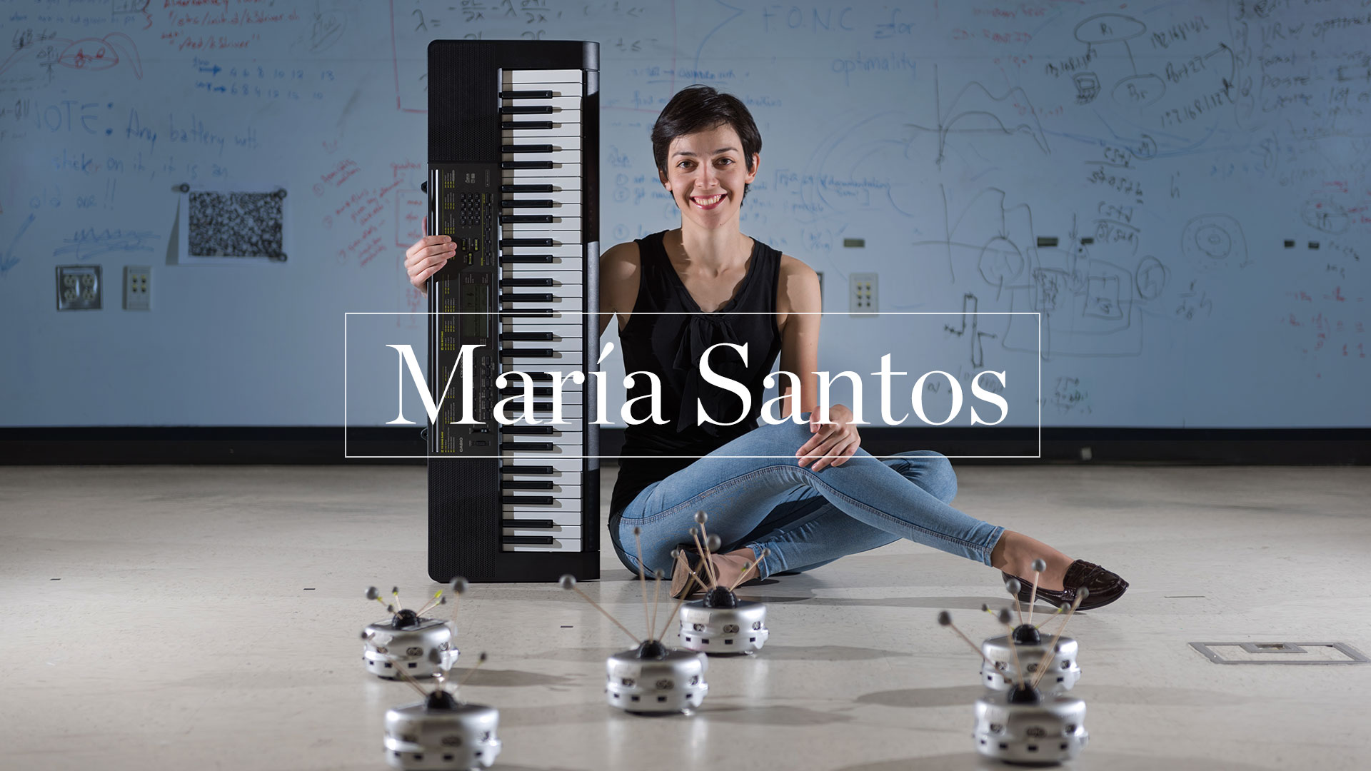 photo -  Maria Santos sitting on floor of computer lab, holding keyboard, with six tiny puck shaped robots on the floor in front of her