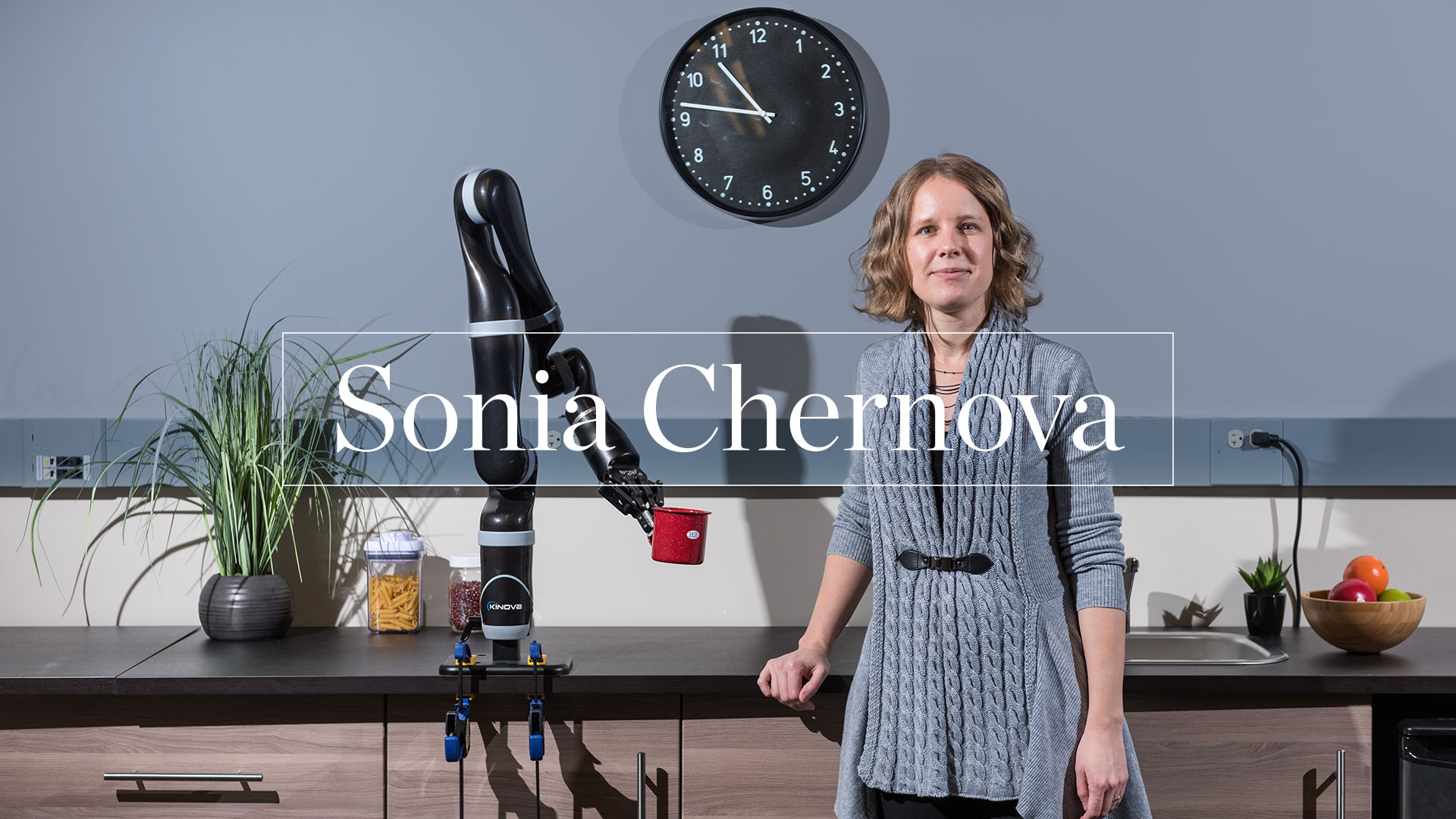 photo -  Sonia Chernova standing in front of counter, with robotic arm mounted to counter top,holding a drink mug