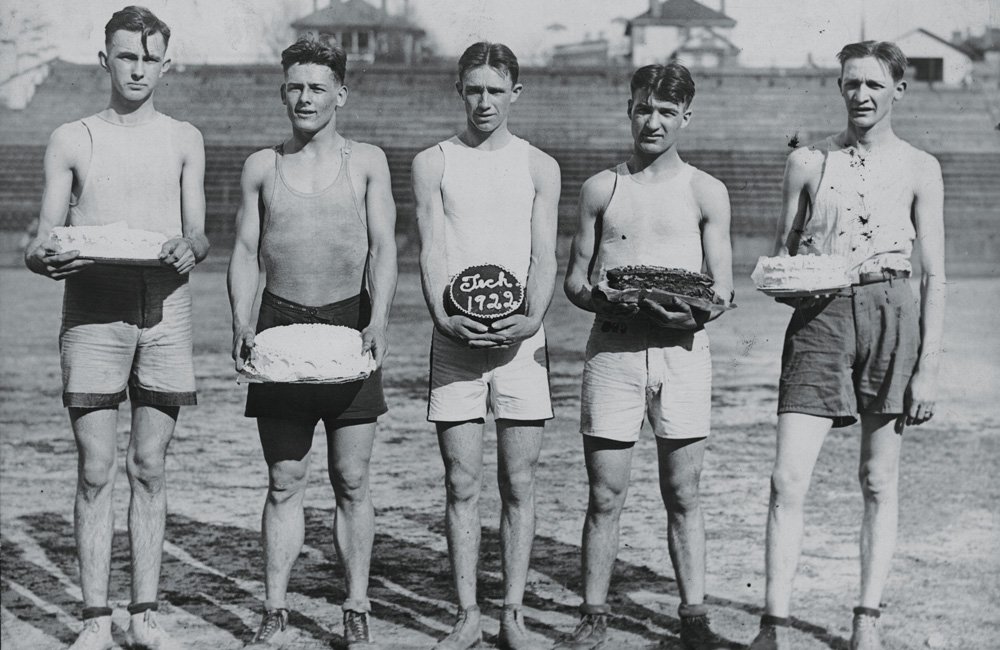 Five young men wearing athletic gear, holding cakes, in a vintage photo