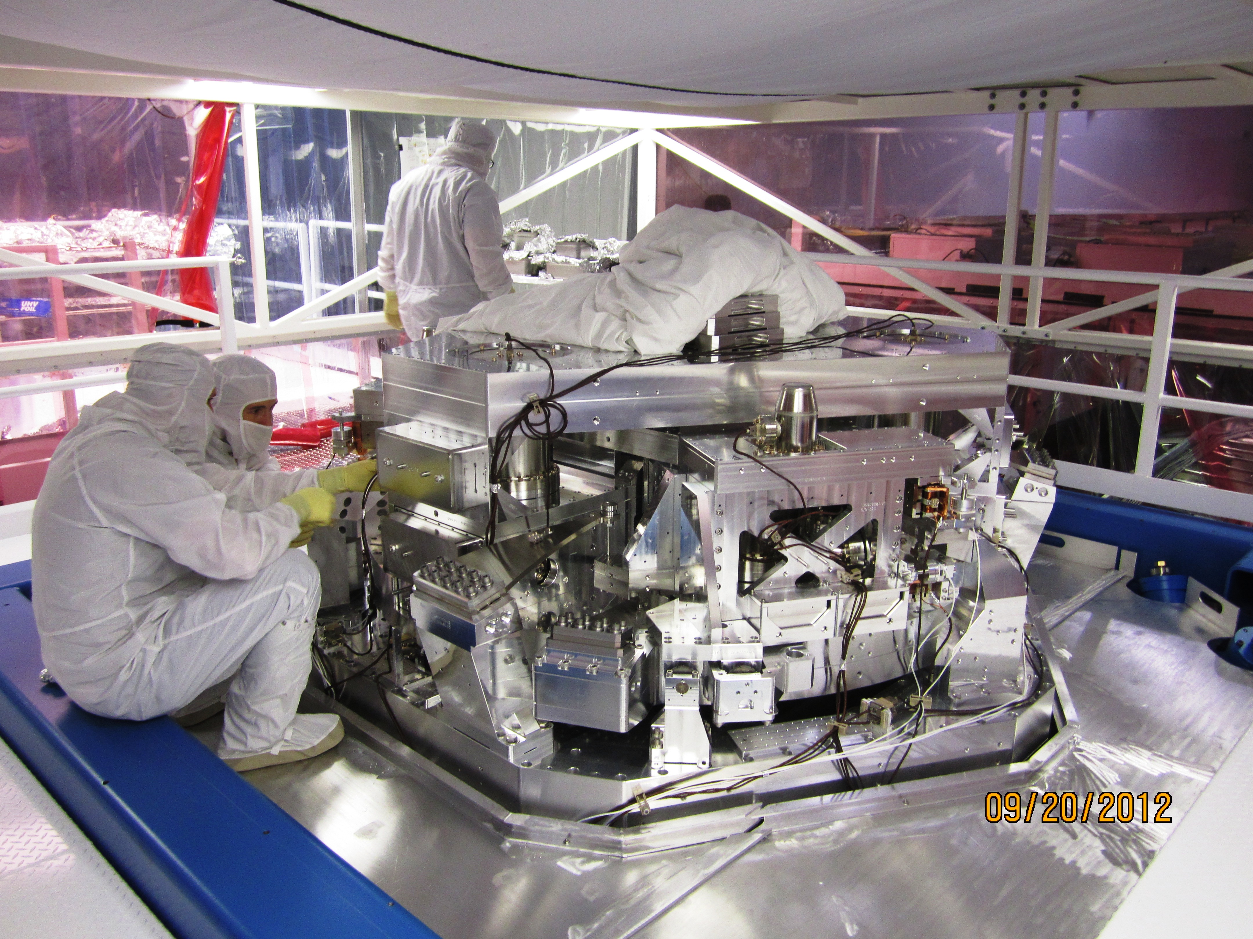 Workers in Livingston, Louisiana are shown installing equipment used to split a powerful laser into two beams, each one traveling down a different arm of the LIGO observatory. The beams bounce off a mirror at the end of each arm and return to the center exactly out of phase, cancelling out the energies of each. (Credit: LIGO Scientific Collaboration)