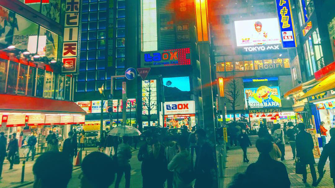 People on the sidewalks in the Akihabara area of Tokyo. The surrounding buildings light the scene with vivid signs.