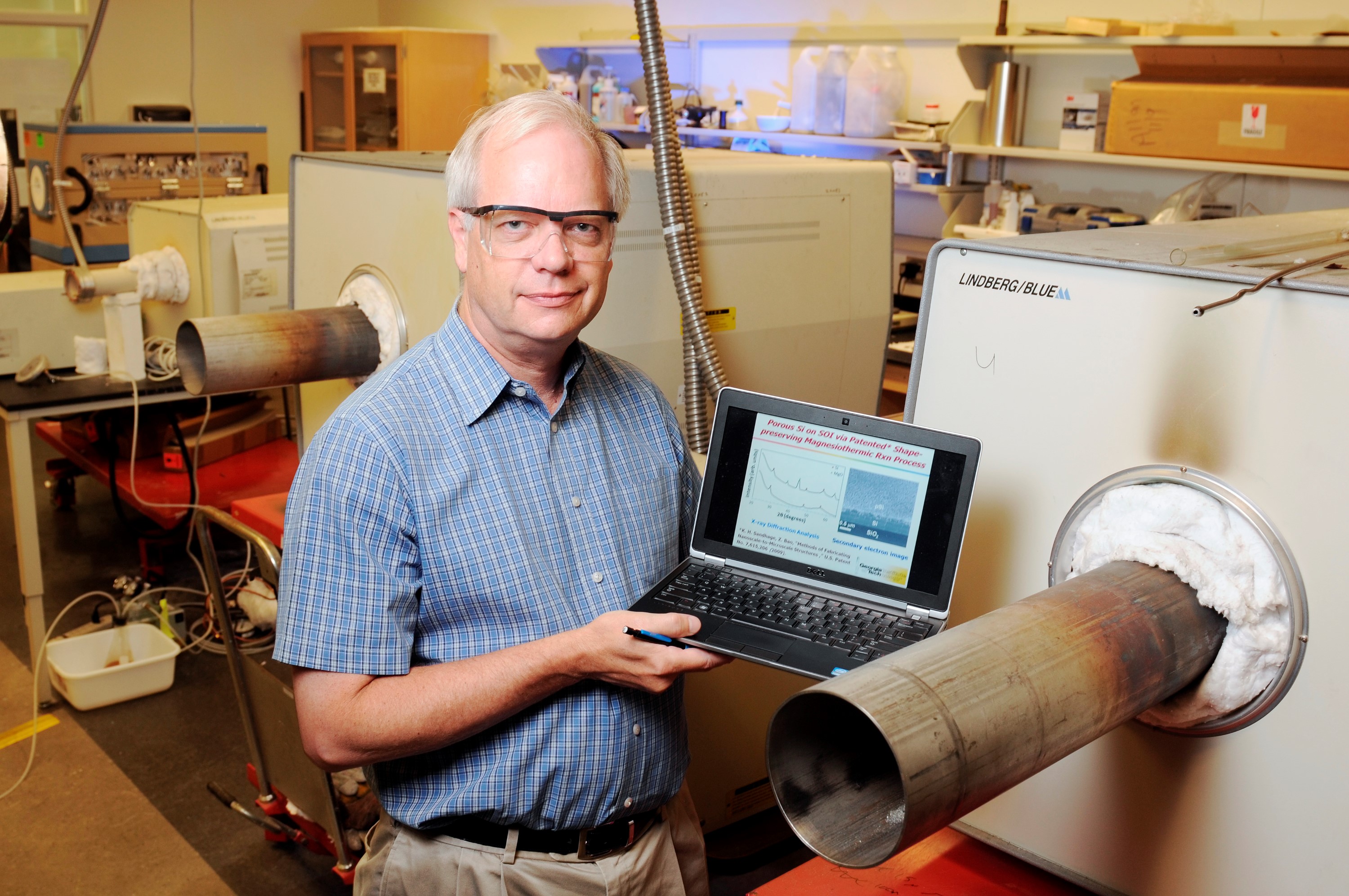 Professor Ken Sandhage, standing near tube furnaces in the Molecular Science and Engineering Building at Georgia Tech, shows a slide demonstrating the ability to generate a porous silicon film on top of a dense silicon layer by selectively reacting silicon dioxide on the dense layer. (Credit: Gary Meek)