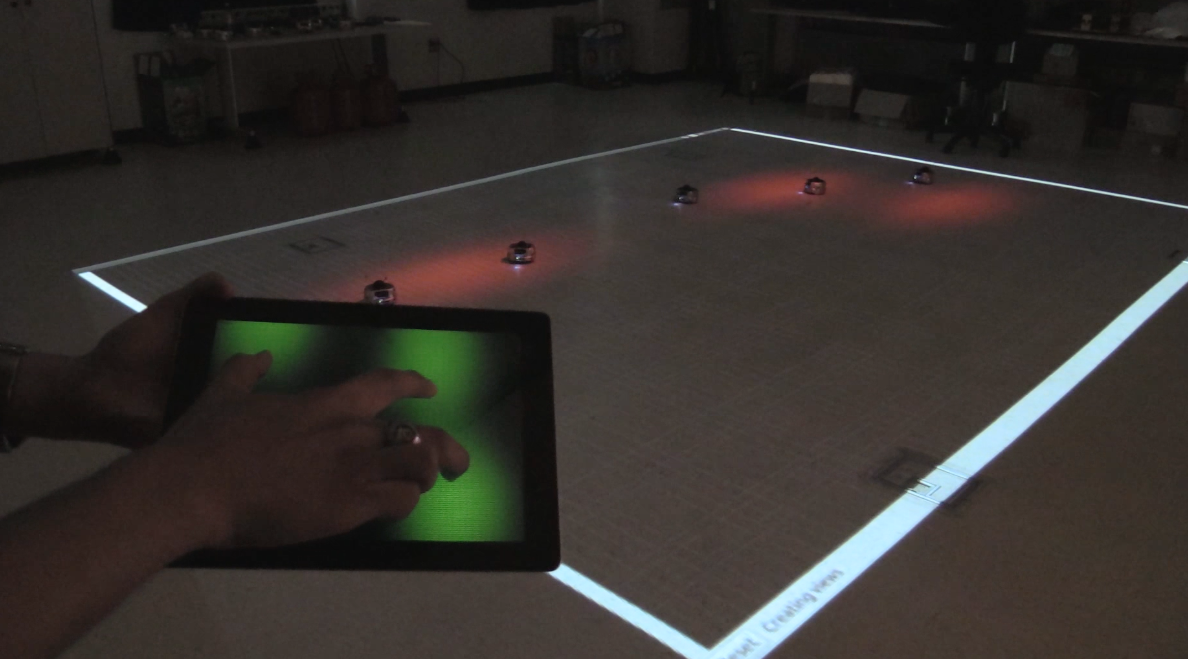 A person taps the tablet to control where the beam of light appears on a floor. The swarm robots then roll toward the illumination, constantly communicating with each other and deciding how to evenly cover the lit area.
