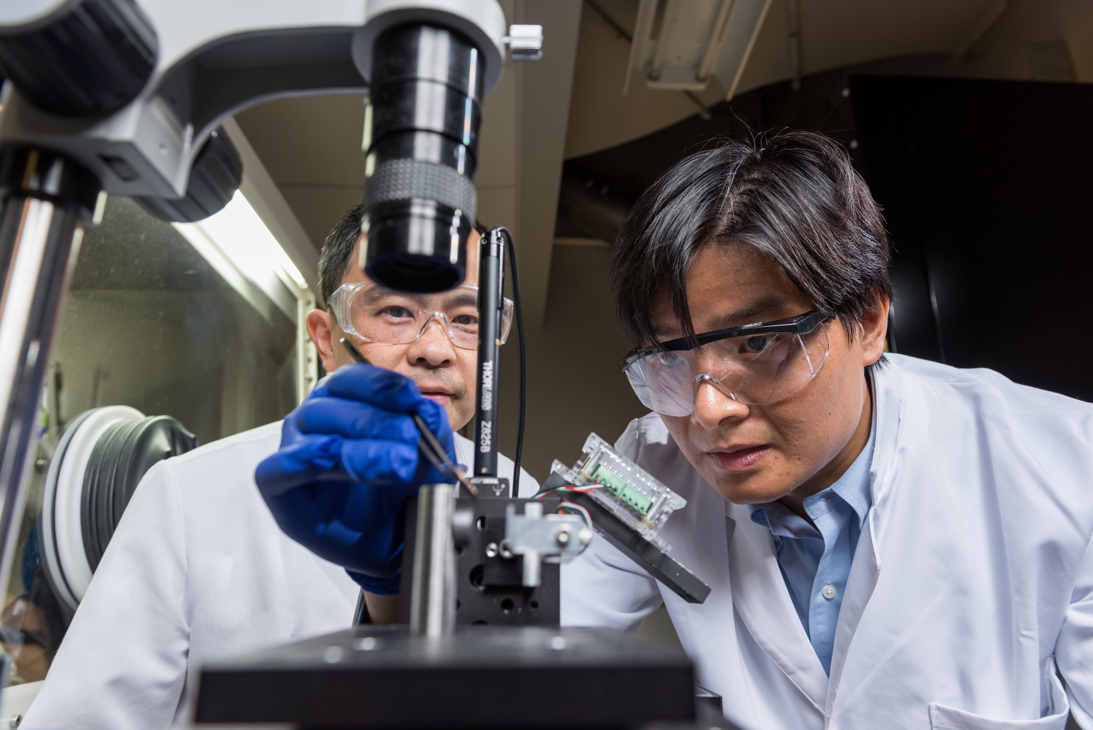 (Left to right) Professor Ting Zhu and Assistant Professor Shuman Xia, both from Georgia Tech’s Woodruff School of Mechanical Engineering, show how a thin film electrode made of amorphous silicon was tested in a custom environmental indenter. To provide proper environmental control, samples containing lithiated silicon were tested with the device inside the glovebox shown in the background. (Credit: Rob Felt, Georgia Tech)