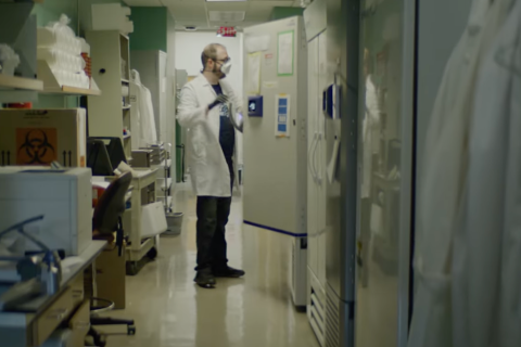 man in lab coat and mask opens freezer to retrieve samples in lab