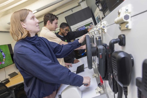 A group of students working on lab equipment. The equipment replicates a ship's helm. 