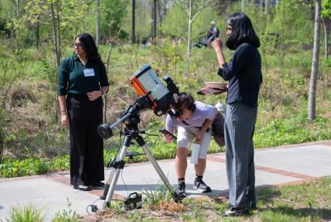 Three people stand outdoors with one person looking at the sun through a telescope