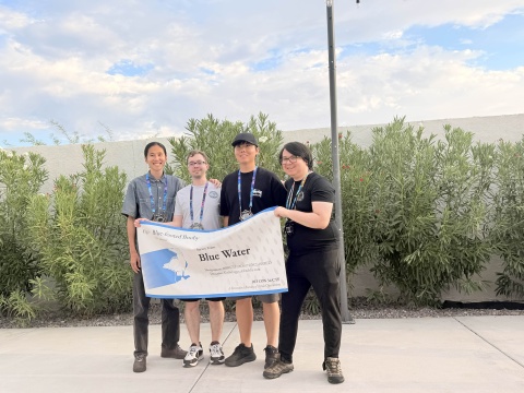 A group of four people stand outside holding up a banner. The banner has their team name, Blue Water