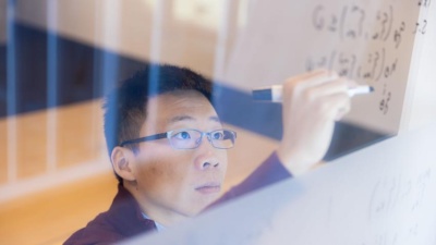 Georgia Tech Assistant Professor Glen Chou with the School of Cybersecurity and Privacy works through an equation on a transparent writing board.