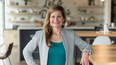 Alison Sizer in a blazer standing in a modern workspace with wooden tables, open shelving, and natural light.