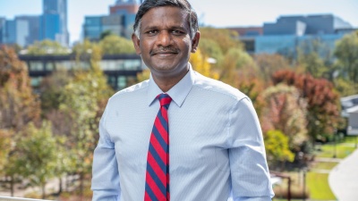 Photo of a man of south Asian descent with short dark hair and a smile. He is wearing a light blue button-down shirt and a red and royal blue striped tie. Behind him is the Atlanta skyline.