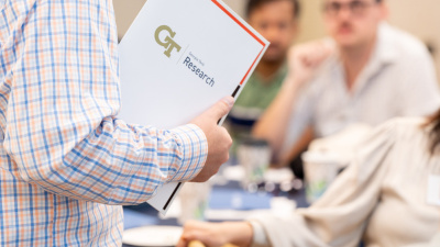 A photo of a new faculty member at orientation, holding a folder that reads "GT Research."