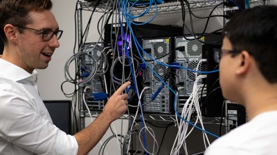 A man points to a rack of CPUs. He is talking to a student who is slighlty out of focus. 