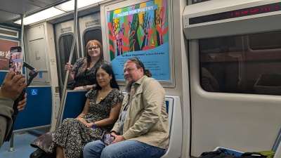 Two people seated in a MARTA train seat underneath a sign with a poem and art in shades of blue, red, purple, green, and black.