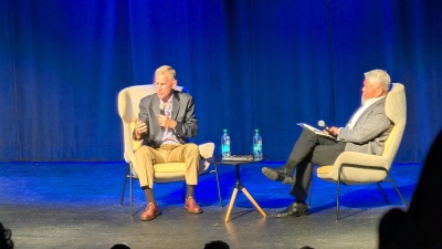 Two men dressed in sportscoats sitting in beige chairs with a blue curtain in the background. A table with water bottles sits between them.
