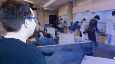 A student wearing safety glasses works in the foreground of a robotics lab as several other students work at equipment in the background.