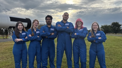 Five people wearing matching blue flight suits stand with arms crossed in a grassy field beside a large rocket display. Sunlight breaks through clouds overhead in the background.
