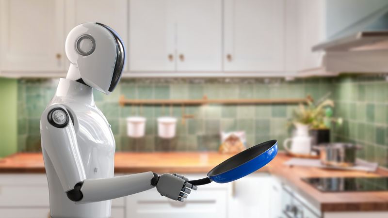 A white humanoid robot holds a blue pan while standing in a kitchen with a green backsplash