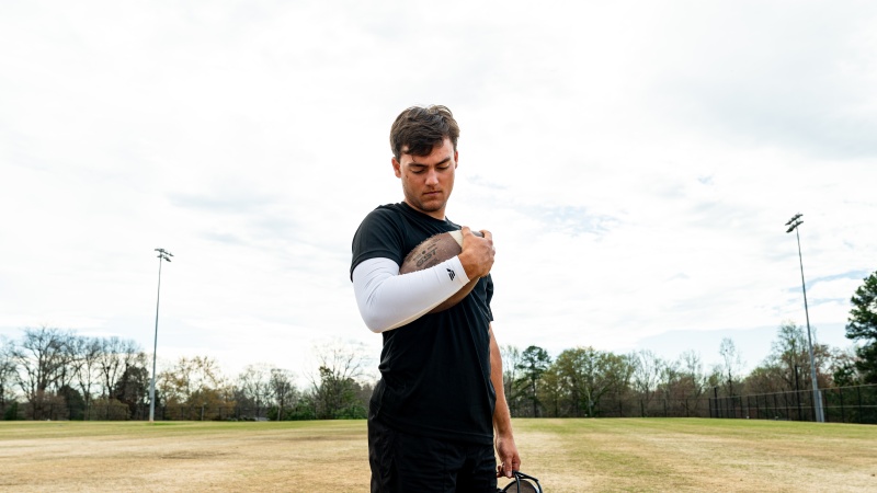 Football player holding a football while wearing a white VETTEX performance arm sleeve on an outdoor field.