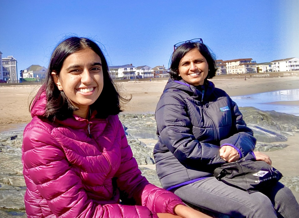 Two smiling researchers wearing jackets sit on rocks at a sunny beach with shoreline homes and the ocean in the background.