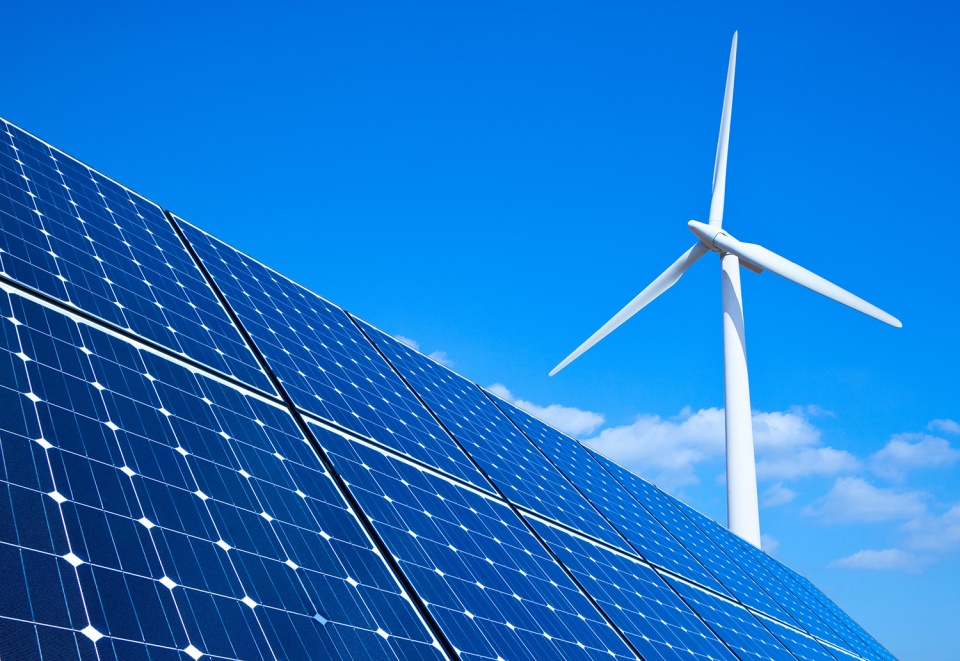 Solar panels cut across the foreground of an image featuring a blue sky and a white wind turbine