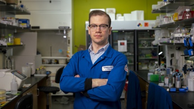A person wearing a blue lab coat stands with arms crossed in a laboratory filled with shelves of scientific equipment, supplies, and a refrigerator unit in the background.