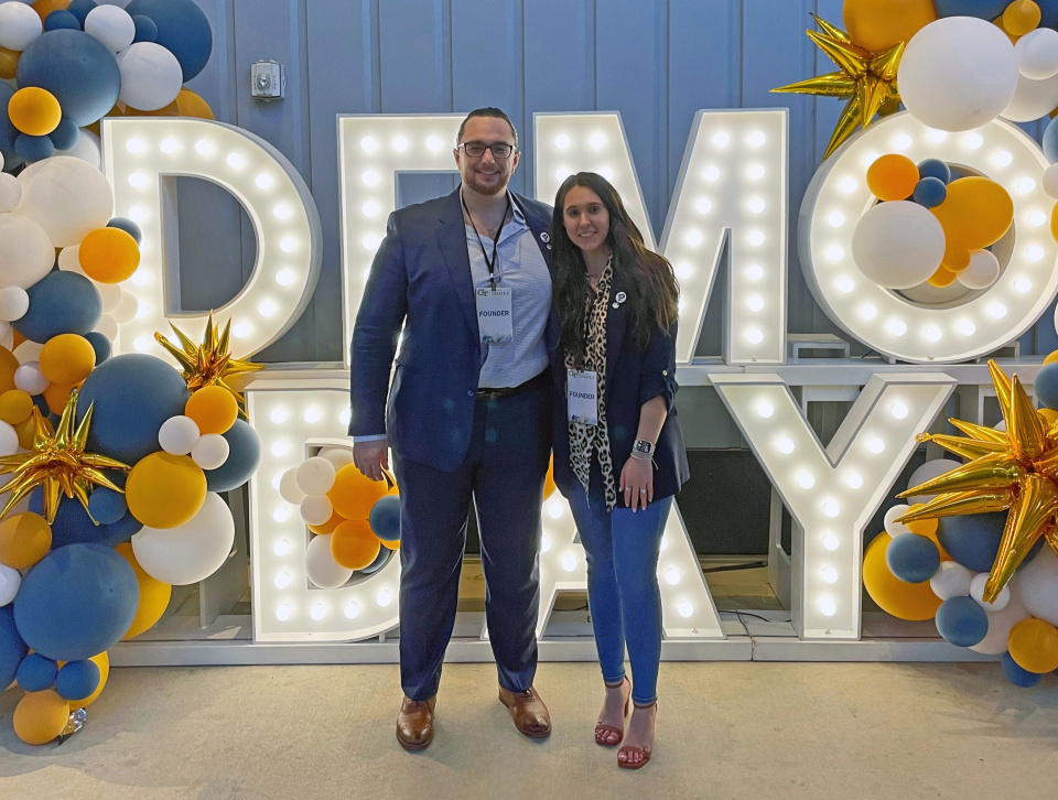 Politheon co-founders Daniel Forcade and Hannah Bodnar stand in front of a lighted sign reading "Demo Day" at the CREATE-X Demo Day in August 2025.