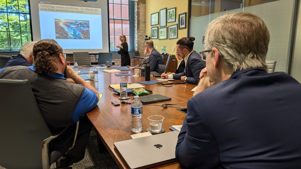 People sitting around a conference table listen to a presentation. A presentation slide reads, "Technology creates possibilities. The liberal arts turns those possibilities into impact."