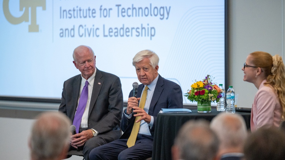 A man speaks into a microphone in front of a banner that reads "Institute for Technology and Civic Leadership" as others look on.
